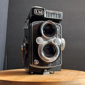 May include: A black YASHICA-MAT LM camera with a silver lens and a Copal-MXV shutter. The camera is on a wooden surface.