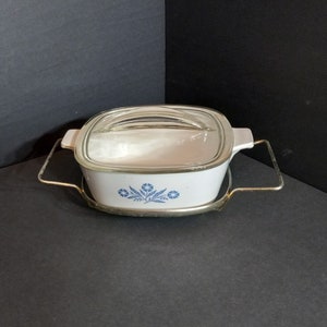 Puede incluir: Plato para hornear blanco con un patr&oacute;n floral azul y una tapa de vidrio. El plato est&aacute; sobre un soporte de metal con asas doradas.