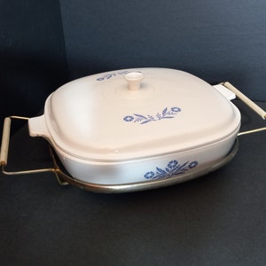 Puede incluir: Plato para hornear cuadrado blanco con un dise&ntilde;o floral azul y una tapa de vidrio. El plato est&aacute; sobre un soporte de metal con dos asas.