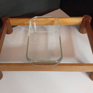 Könnte beinhalten: Ein Servierständer aus Holz mit vier abgerundeten Beinen und einem rechteckigen Rahmen. Eine klare Glasbackform sitzt auf dem Rahmen.