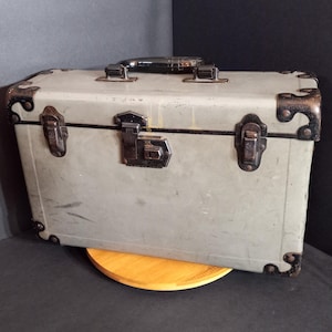 May include: A vintage gray trunk with dark metal hardware, including corner protectors, latches, and a handle. The trunk is rectangular and sits on a wooden stand. The metal hardware has a dark, aged appearance.