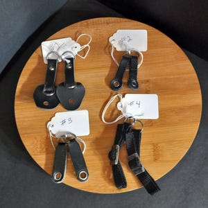 May include: Four sets of black straps with metal hardware and white paper tags labeled #1, #2, #3, and #4. The straps vary in shape, including rounded and rectangular ends with metal grommets or rings, arranged on a round wooden surface.