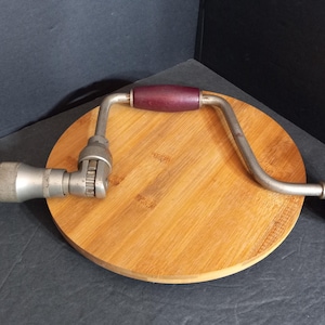 Puede incluir: Un taladro manual vintage con un cuerpo de metal plateado, un mango de madera rojo oscuro y una broca. El taladro está sobre una superficie redonda de madera marrón claro. El fondo es oscuro.