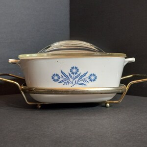 Puede incluir: Una fuente para horno blanca con un dise&ntilde;o floral azul y una tapa de vidrio, sentada sobre un soporte de metal dorado.