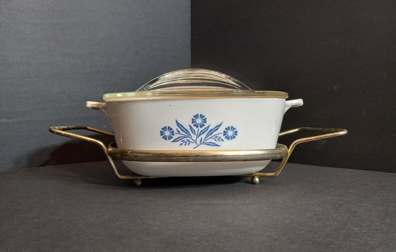 Puede incluir: Una fuente para horno blanca con un dise&ntilde;o floral azul y una tapa de vidrio, sentada sobre un soporte de metal dorado.