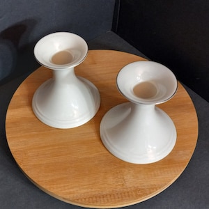 May include: Two white ceramic candle holders with a silver rim on a wooden surface. The candle holders are shaped like a wide bowl with a narrow base.