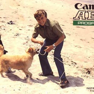 May include: A vintage photograph shows a person on a beach with two dogs. The person is holding a leash, and one dog is biting it. The image has text that reads "Canon AE-1 PROGRAM" and "INSTRUCTIONS English Edition."