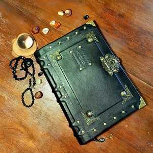 Puede incluir: Un diario encuadernado en cuero negro con detalles dorados y una cerradura de lat&oacute;n. El diario tiene un dise&ntilde;o decorativo en la portada y la palabra "AOO" est&aacute; grabada en relieve en la parte delantera.