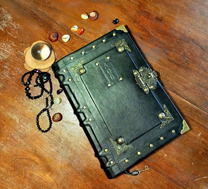 Puede incluir: Un diario encuadernado en cuero negro con detalles dorados y una cerradura de lat&oacute;n. El diario tiene un dise&ntilde;o decorativo en la portada y la palabra "AOO" est&aacute; grabada en relieve en la parte delantera.