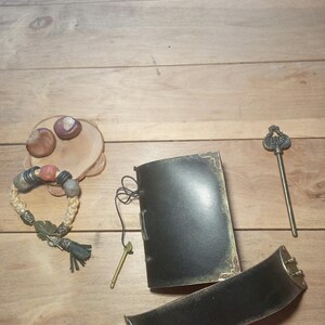May include: An assortment of handcrafted items on a wooden surface. Includes a black leather journal with gold trim, a woven bracelet with beads, a decorative key, and a wide black belt with a brass buckle. Also, a small wooden plate with two decorative stones.