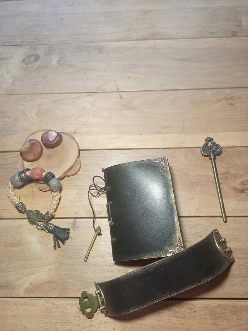 May include: An assortment of handcrafted items on a wooden surface. Includes a black leather journal with gold trim, a woven bracelet with beads, a decorative key, and a wide black belt with a brass buckle. Also, a small wooden plate with two decorative stones.
