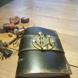 May include: A black leather journal with a brass clasp and corner accents. The journal is bound with a wide, metallic gold band. A decorative beaded tassel and two chestnuts are visible in the background. The journal is on a wooden surface.