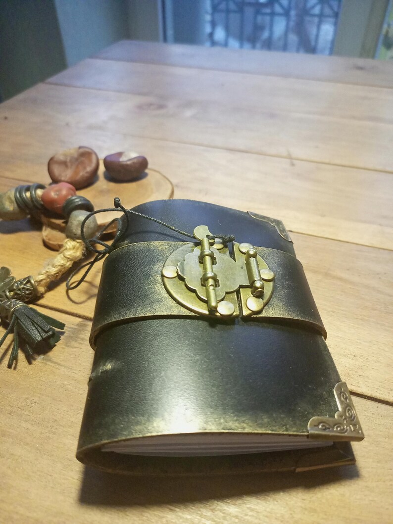 May include: A black leather journal with a brass clasp and corner accents. The journal is bound with a wide, metallic gold band. A decorative beaded tassel and two chestnuts are visible in the background. The journal is on a wooden surface.