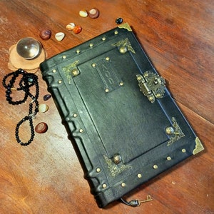 Puede incluir: Un diario encuadernado en cuero negro con detalles dorados y una cerradura de lat&oacute;n. El diario tiene un aspecto desgastado y est&aacute; decorado con detalles met&aacute;licos decorativos.