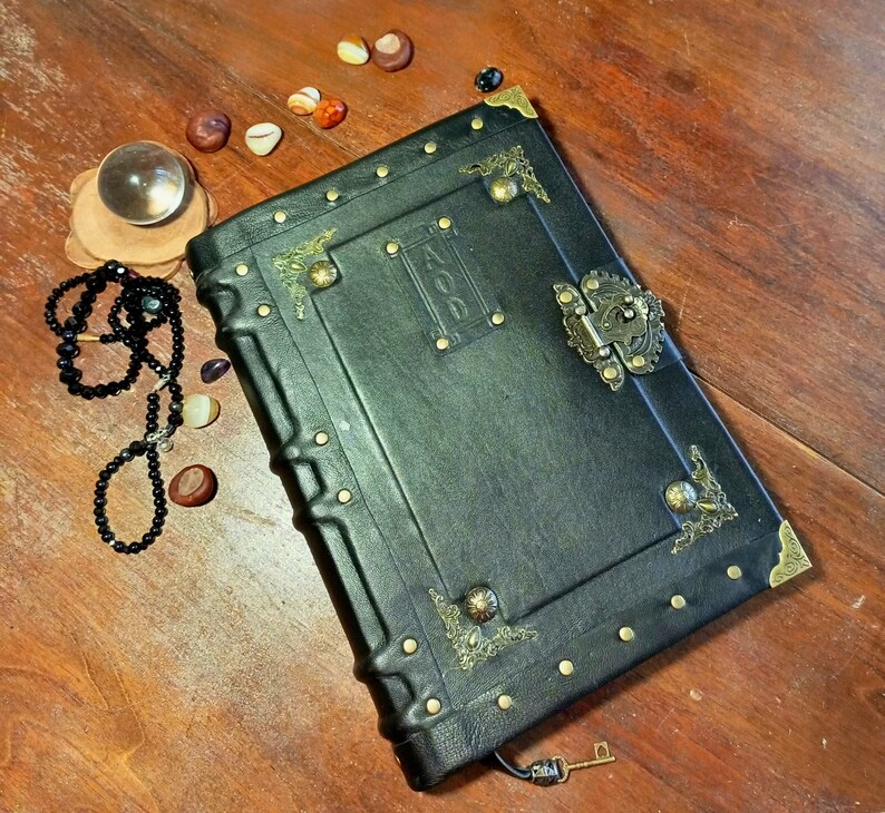 Puede incluir: Un diario encuadernado en cuero negro con detalles dorados y una cerradura de lat&oacute;n. El diario tiene un aspecto desgastado y est&aacute; decorado con detalles met&aacute;licos decorativos.