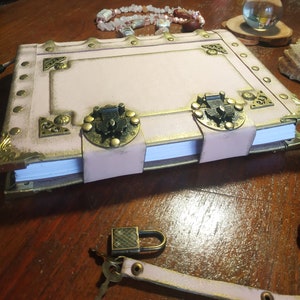 May include: A pink leather-bound journal with ornate brass accents and clasps. The journal has a white interior and is closed. A small padlock is visible in the foreground.