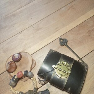 May include: An assortment of handcrafted items on a wooden surface. Includes a black leather journal with a brass clasp, a beaded bracelet with a leaf charm, a decorative key, and a small wooden dish with chestnuts.