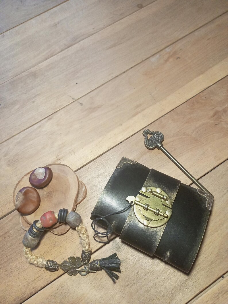 May include: An assortment of handcrafted items on a wooden surface. Includes a black leather journal with a brass clasp, a beaded bracelet with a leaf charm, a decorative key, and a small wooden dish with chestnuts.
