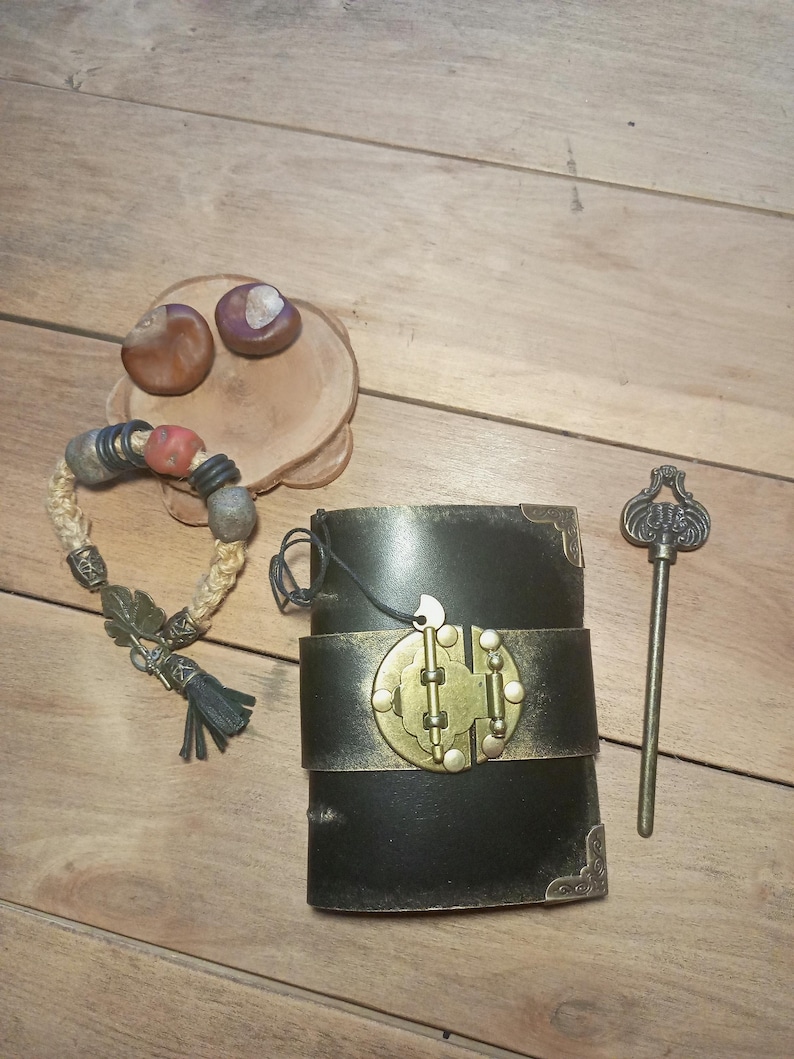 May include: A black leather journal with a brass clasp and a decorative key. A beaded bracelet with a tassel and a small wooden plate with two chestnuts are also present. The items are arranged on a wooden surface.