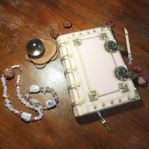 May include: A pink leather-bound journal with gold accents and brass clasps. The journal has a keyhole and a key attached to a leather strap. The journal is on a wooden surface with a crystal ball, a necklace, and some chestnuts.