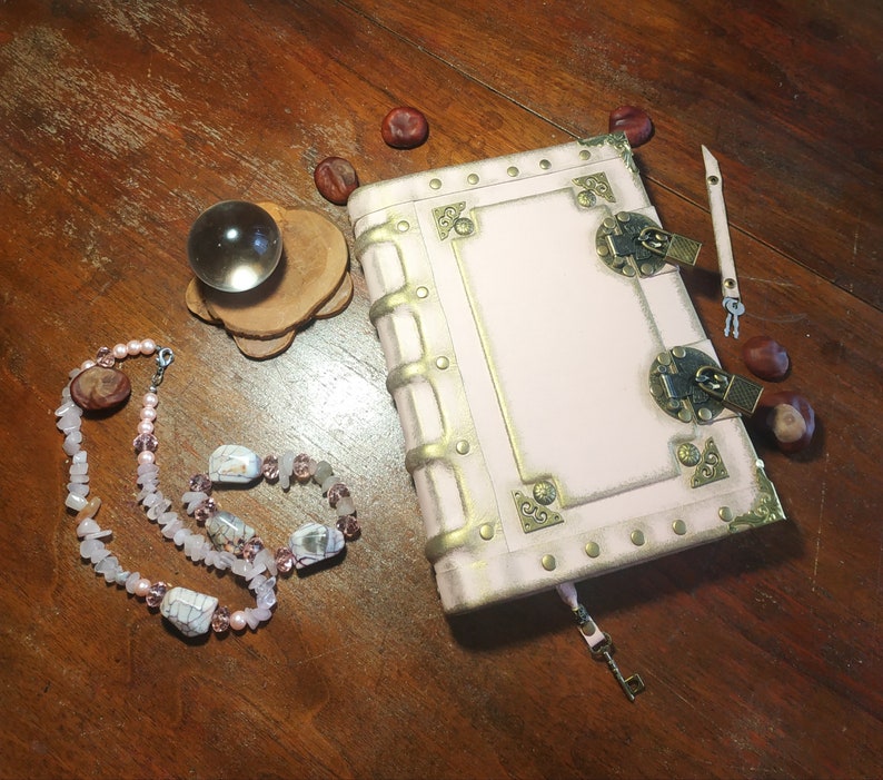 May include: A pink leather-bound journal with gold accents and brass clasps. The journal has a keyhole and a key attached to a leather strap. The journal is on a wooden surface with a crystal ball, a necklace, and some chestnuts.