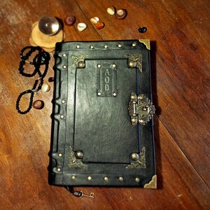 Puede incluir: Un diario encuadernado en cuero negro con detalles dorados y una cerradura de lat&oacute;n. El diario tiene un dise&ntilde;o decorativo y la palabra "AOD" grabada en la portada.