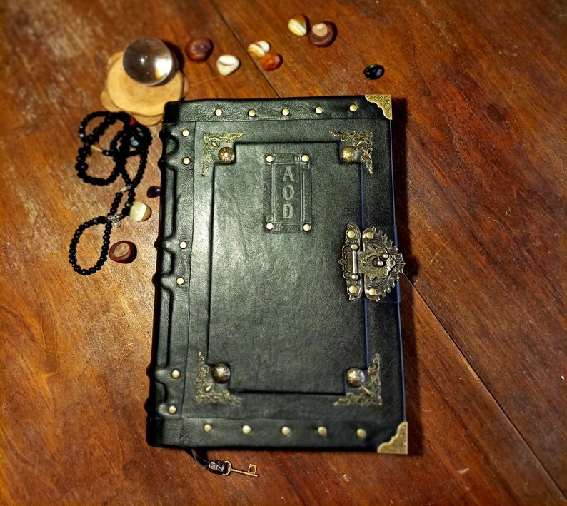 Puede incluir: Un diario encuadernado en cuero negro con detalles dorados y una cerradura de lat&oacute;n. El diario tiene un dise&ntilde;o decorativo y la palabra "AOD" grabada en la portada.