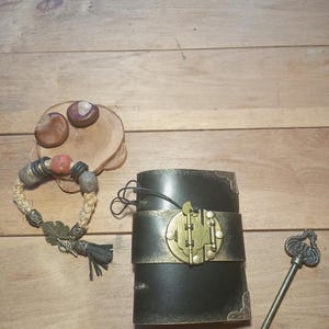 May include: A black leather journal with a brass clasp and a decorative key. A braided bracelet with beads and a tassel is next to the journal. Two chestnuts sit on a wooden slice.