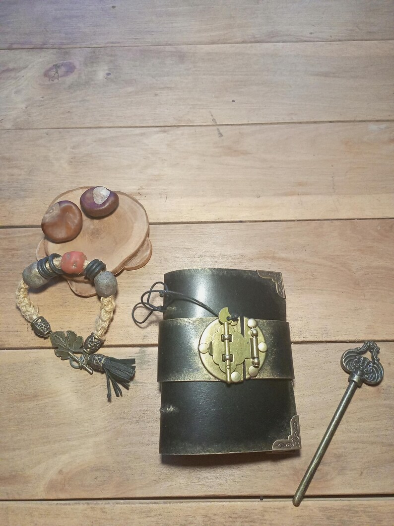 May include: A black leather journal with a brass clasp and a decorative key. A braided bracelet with beads and a tassel is next to the journal. Two chestnuts sit on a wooden slice.