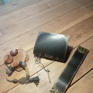 May include: A collection of handcrafted leather and beaded accessories. Includes a dark leather journal, a wide leather bracelet with a brass clasp, and a beaded bracelet with a leaf charm and tassel.