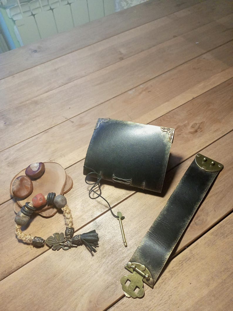 May include: A collection of handcrafted leather and beaded accessories. Includes a dark leather journal, a wide leather bracelet with a brass clasp, and a beaded bracelet with a leaf charm and tassel.
