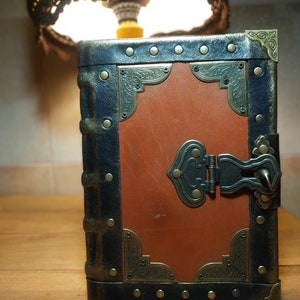 May include: A brown leather-bound journal with a black metal frame, brass accents, and a metal clasp. The journal has a tassel attached to the bottom.