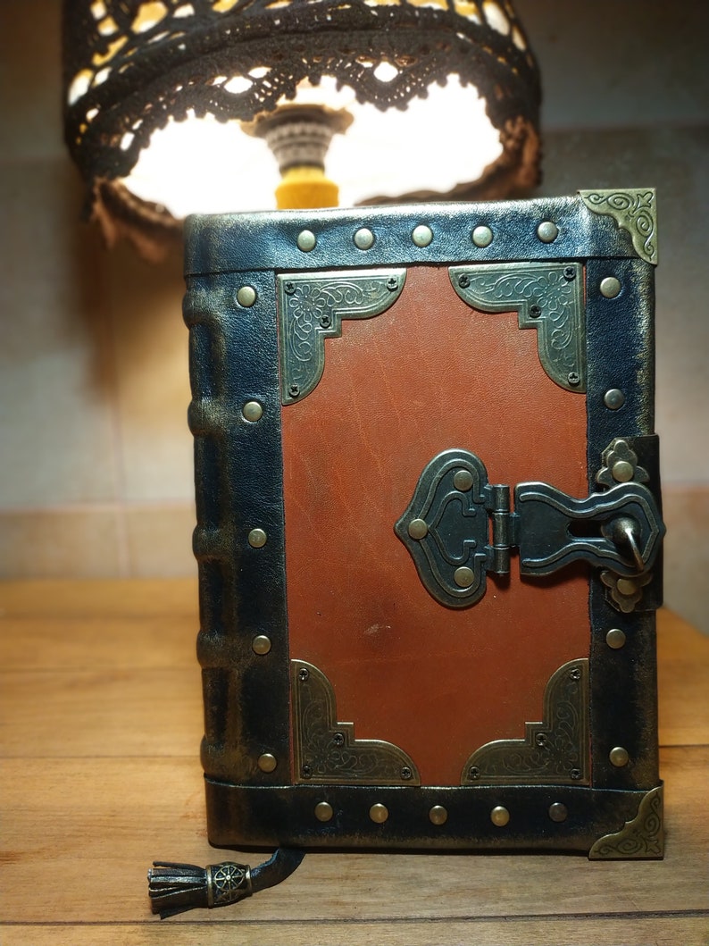 May include: A brown leather-bound journal with a black metal frame, brass accents, and a metal clasp. The journal has a tassel attached to the bottom.