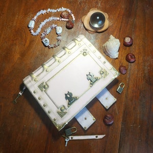 May include: A white leather-bound journal with gold accents and decorative metal clasps. The journal is closed and lying on a wooden surface with a necklace and other decorative items.