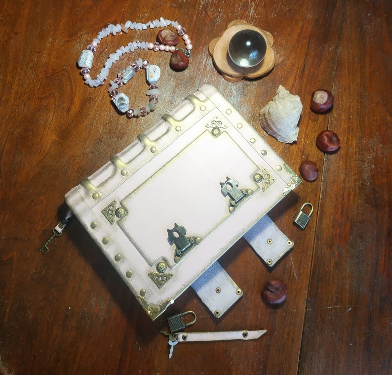 May include: A white leather-bound journal with gold accents and decorative metal clasps. The journal is closed and lying on a wooden surface with a necklace and other decorative items.