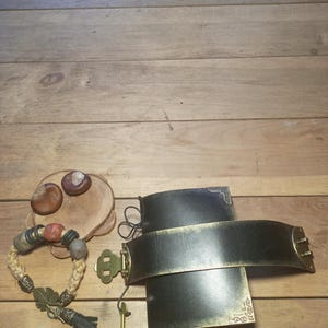 May include: A collection of handcrafted items on a wooden surface. Includes a black leather cuff with brass accents, a beaded bracelet with natural stone beads, and a small wooden coaster with chestnuts.