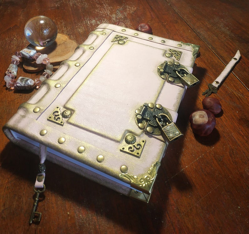 May include: A pink leather-bound journal with gold accents and two brass locks. The journal has a key attached to it and is sitting on a wooden surface.