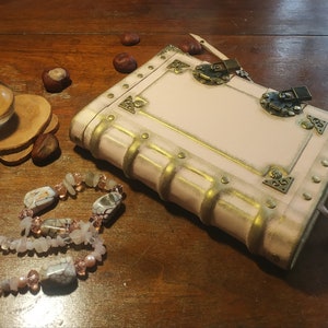 May include: A pink leather-bound journal with gold accents and a metal clasp. The journal has a keyhole and a key attached to it. The journal is sitting on a wooden surface with a crystal ball and a necklace of pink and white stones.