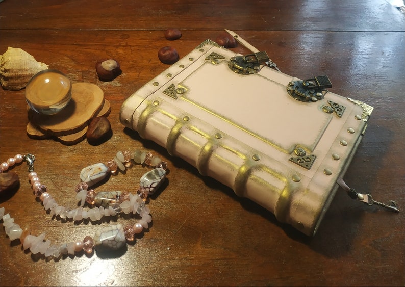 May include: A pink leather-bound journal with gold accents and a metal clasp. The journal has a keyhole and a key attached to it. The journal is sitting on a wooden surface with a crystal ball and a necklace of pink and white stones.