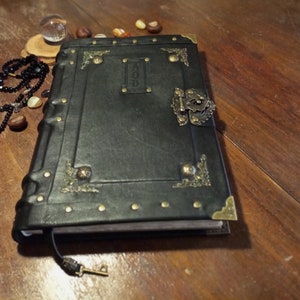 Puede incluir: Un diario encuadernado en cuero negro con detalles en lat&oacute;n y un cierre de metal. El diario tiene un dise&ntilde;o decorativo en la portada y la palabra "AOD" est&aacute; grabada en la portada.