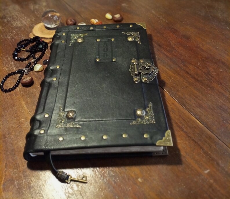 Puede incluir: Un diario encuadernado en cuero negro con detalles en lat&oacute;n y un cierre de metal. El diario tiene un dise&ntilde;o decorativo en la portada y la palabra "AOD" est&aacute; grabada en la portada.