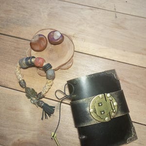 May include: A black leather cuff bracelet with a gold-colored clasp and accents, next to a beaded bracelet with a tassel and a small key. Two chestnuts rest on a wooden coaster.