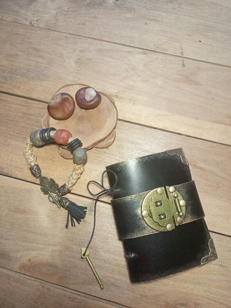 May include: A black leather cuff bracelet with a gold-colored clasp and accents, next to a beaded bracelet with a tassel and a small key. Two chestnuts rest on a wooden coaster.