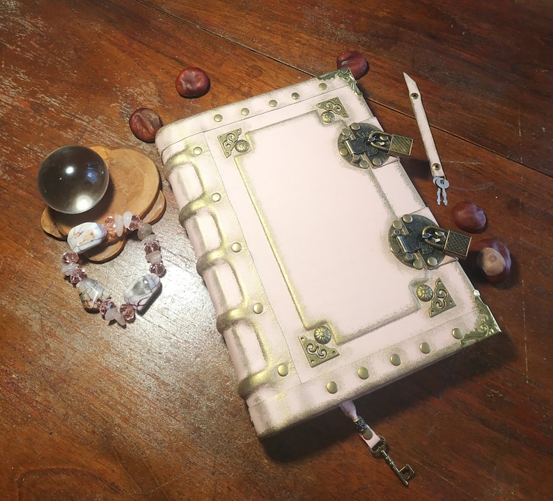 May include: A pink leather-bound journal with gold accents and two brass clasps. The journal has a distressed look and is decorated with rivets and decorative metal pieces.