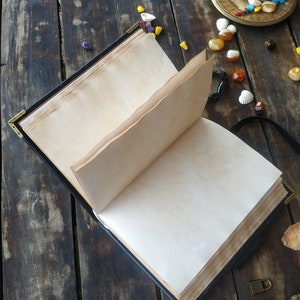 May include: An open black leather-bound journal with blank pages, lying on a dark wooden surface. There are small colorful stones and seashells scattered around the journal, along with a candle holder and a key.