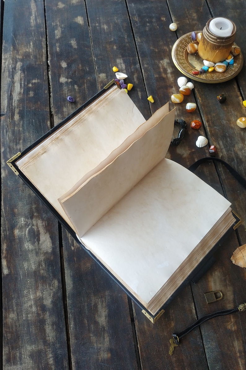 May include: An open black leather-bound journal with blank pages, lying on a dark wooden surface. There are small colorful stones and seashells scattered around the journal, along with a candle holder and a key.