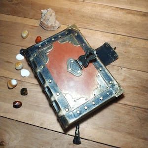 May include: A dark brown leather journal with a metal clasp and decorative accents. The journal has a distressed look and is adorned with gold and brass accents.