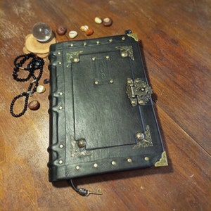 Puede incluir: Un diario encuadernado en cuero negro con detalles dorados y un cierre de metal. El diario tiene una cerradura y una peque&ntilde;a llave adjunta. El diario est&aacute; sobre una superficie de madera.