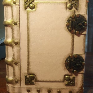 May include: A white leather-bound journal with gold accents and brass clasps. The journal has a distressed look and is decorated with decorative metal corners.