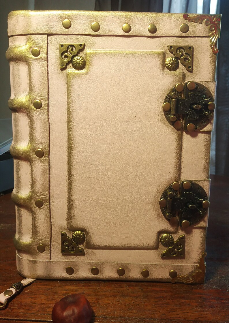 May include: A white leather-bound journal with gold accents and brass clasps. The journal has a distressed look and is decorated with decorative metal corners.